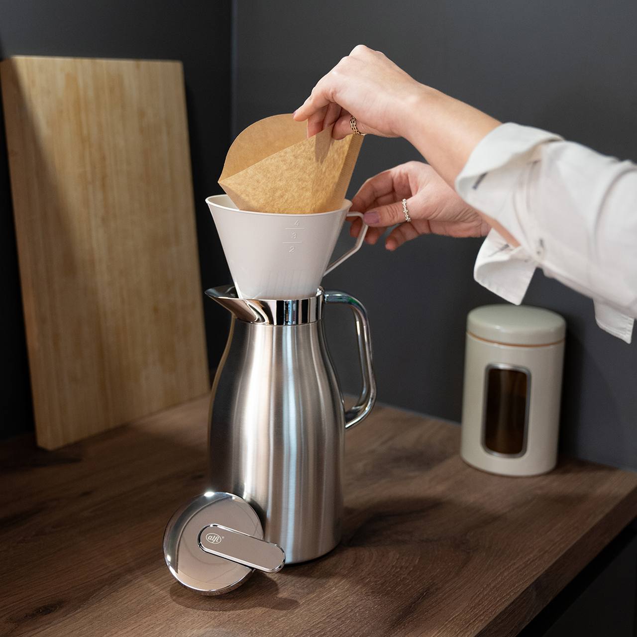 Jemand bereitet Kaffee mit einem silbernen Filterhalter und einer Glaskanne vor und legt einen braunen Kaffeefilter ein. Daneben stehen ein Holzbrett an der Wand und ein weißer Behälter mit Schriftzug. Die Szene ist in einer modernen Küche.