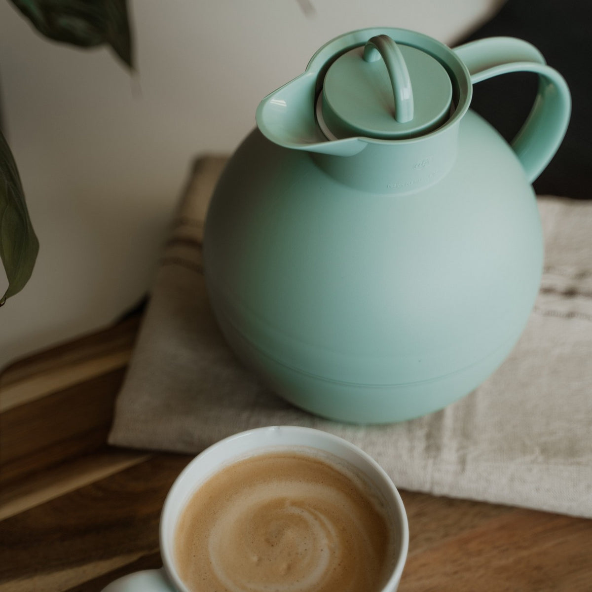 Auf dem Bild sieht man eine mintgrüne Thermoskanne, die auf einem beigen Tuch steht. Daneben steht eine Tasse, die mit einem cremigen Cappuccino gefüllt ist. Die Szene wirkt ruhig und einladend.