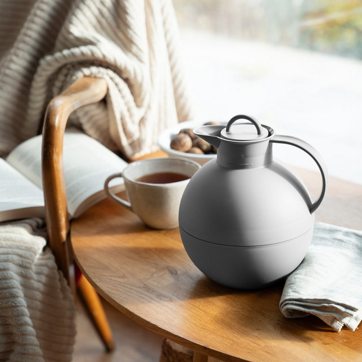 Auf einem Tisch vor einem Fenster stehen eine graue Thermoskanne, eine Tasse Tee und ein gefaltetes Tuch. Daneben auf einem Stuhl liegen eine Decke und ein aufgeschlagenes Buch. Im Hintergrund sind Nüsse auf einem Teller.