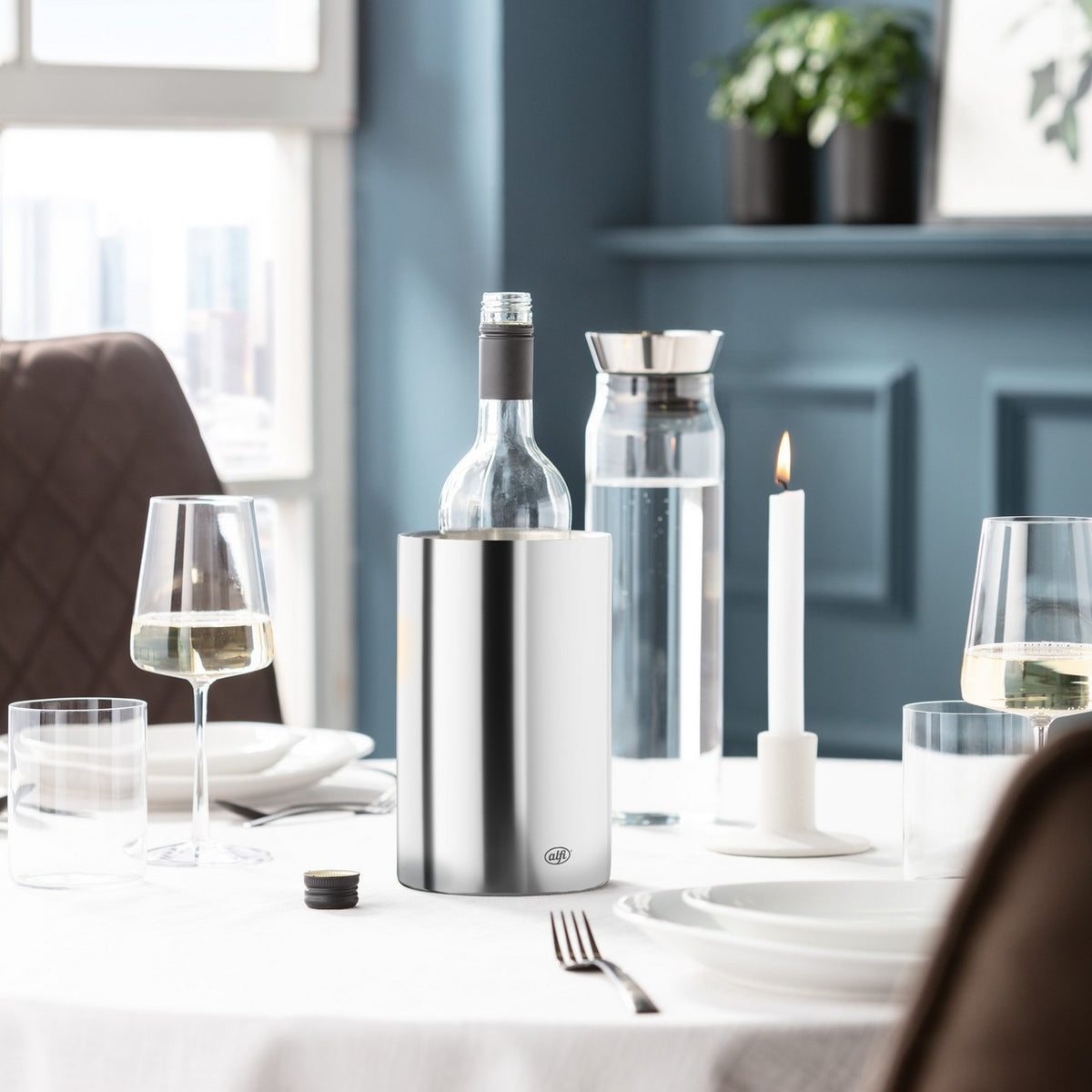 Das Bild zeigt einen elegant gedeckten Tisch mit weißen Kerzen in Kerzenhaltern und einer Edelstahl-Isolierkanne. Im Hintergrund ist eine blaue Wand mit einem Regal, auf dem Pflanzen stehen. Durch das Fenster fällt Tageslicht in den Raum.