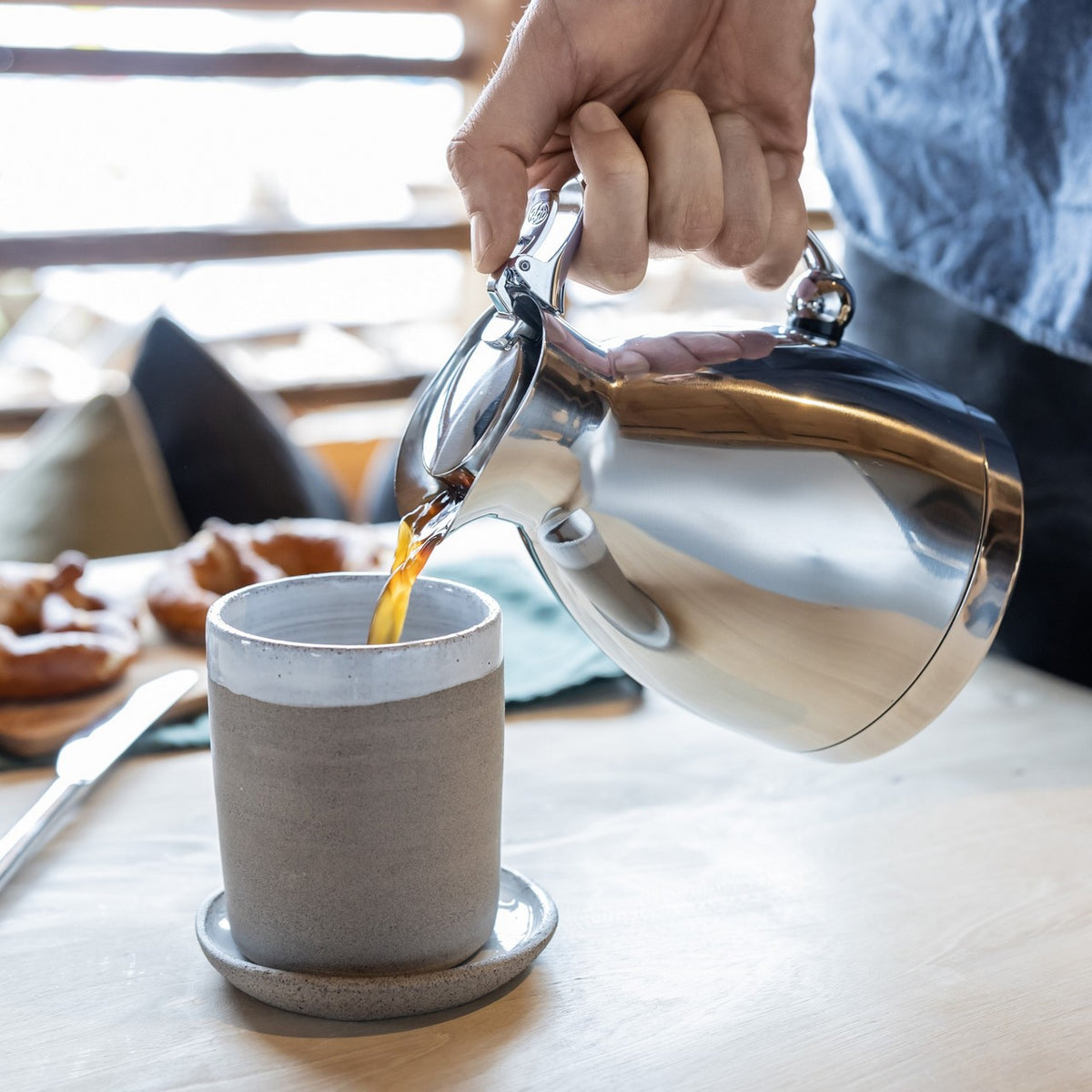 Eine Person gießt Kaffee aus einer silbernen Kanne in eine graue Tasse. Im Hintergrund sind verschwommen Snacks wie Schokokekse zu sehen. Der Tisch hat eine helle Holzoberfläche.