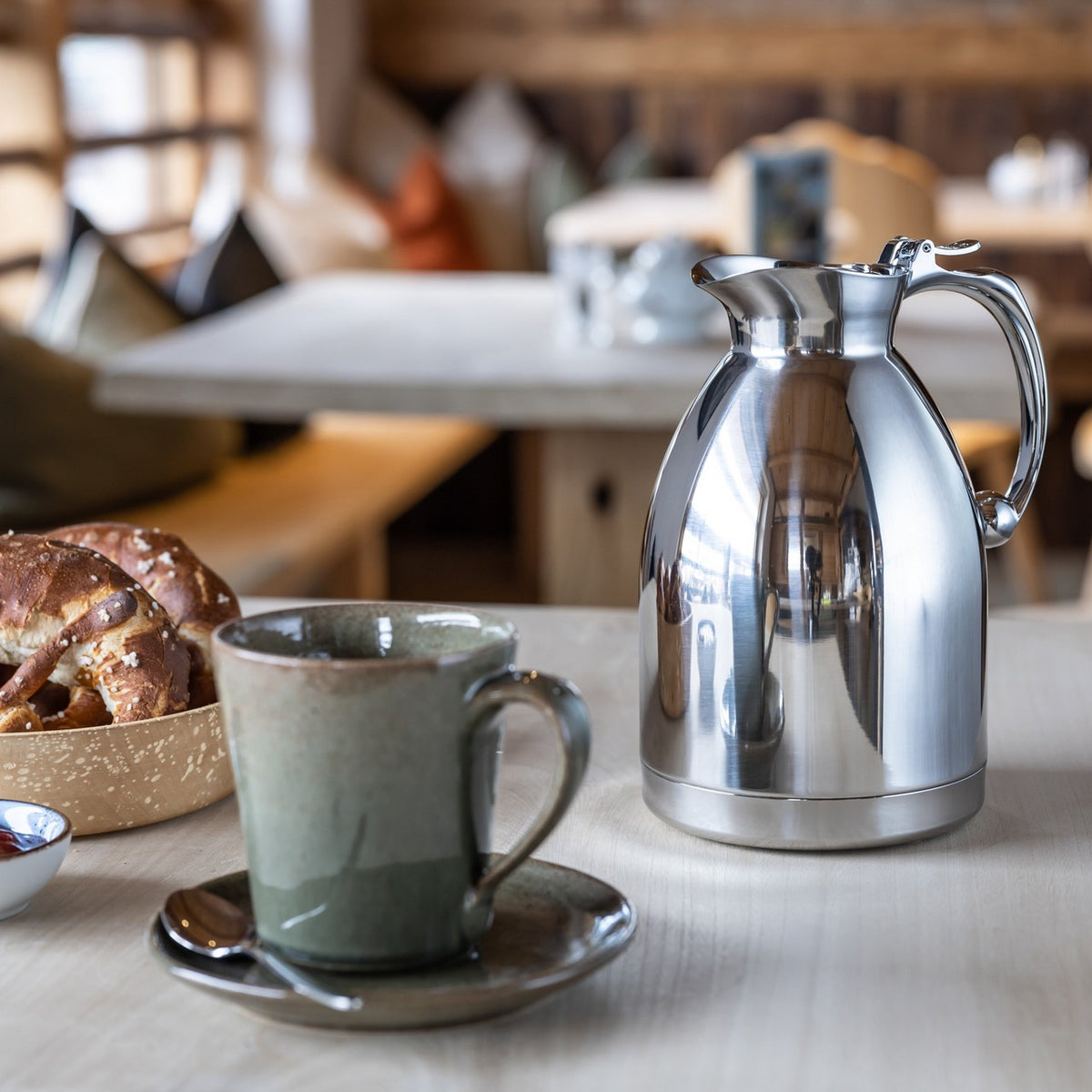 Auf dem Bild befindet sich eine graue Tasse mit Kaffee auf einer Untertasse, daneben stehen zwei silberne Thermoskannen. Im Hintergrund ist ein gemütlicher Raum mit Holzmöbeln und dekorativen Gegenständen zu sehen.