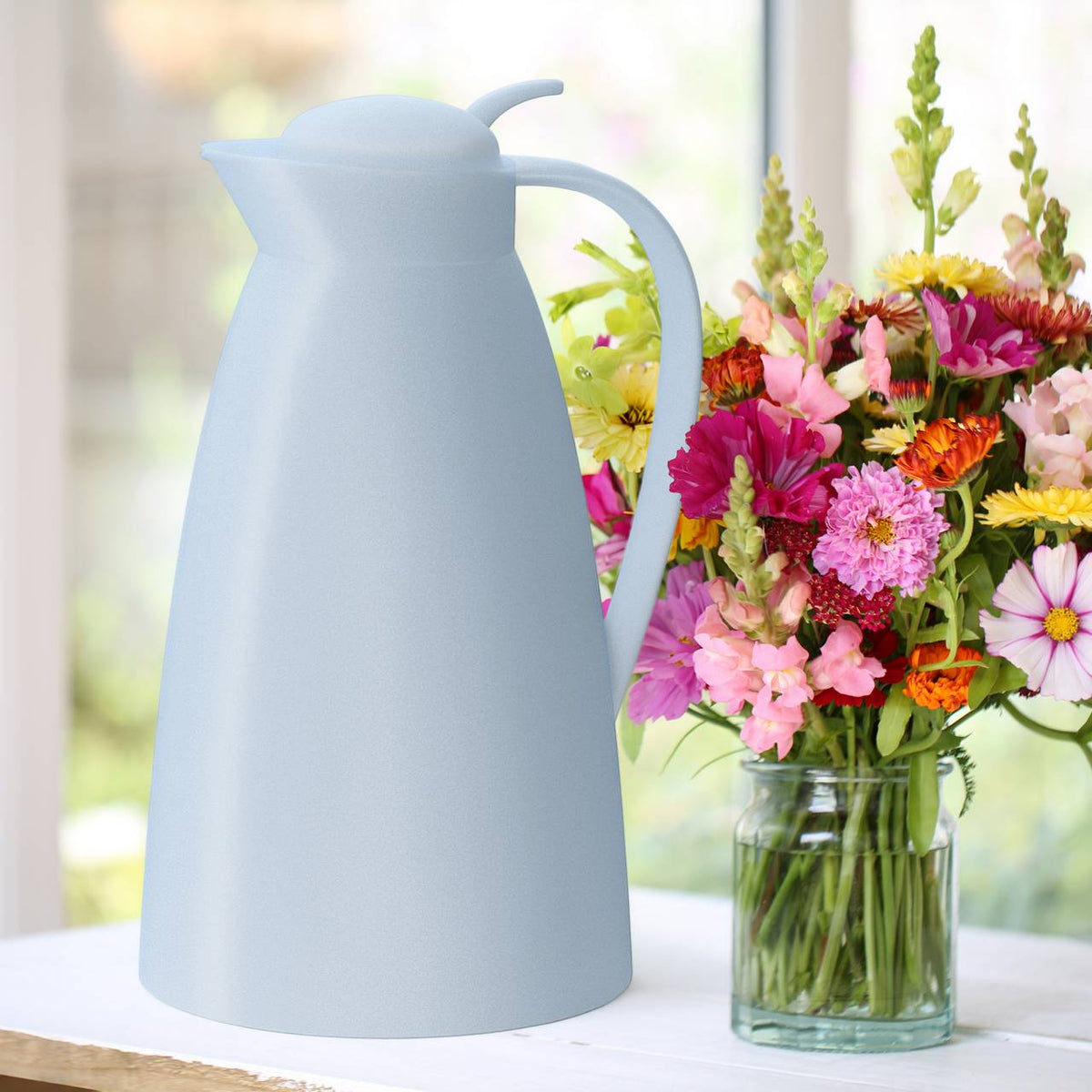 Auf dem Bild steht eine hellblaue Thermoskanne neben einem Strauß bunter Blumen in einer Glasvase. Beide Objekte befinden sich auf einem weißen Tisch vor einem unscharfen Hintergrund, der auf ein Interieur mit viel natürlichem Licht schließen lässt.