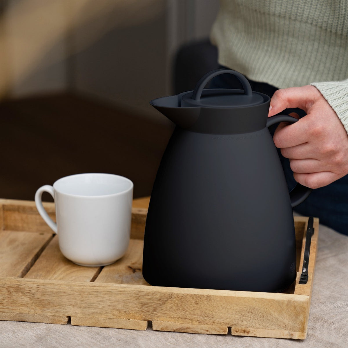 Eine Person hält eine schwarze Kanne auf einem Holztablett, das auch eine weiße Tasse enthält. Das Tablett steht auf einem Tisch, und die Person trägt einen hellen Strickpullover.