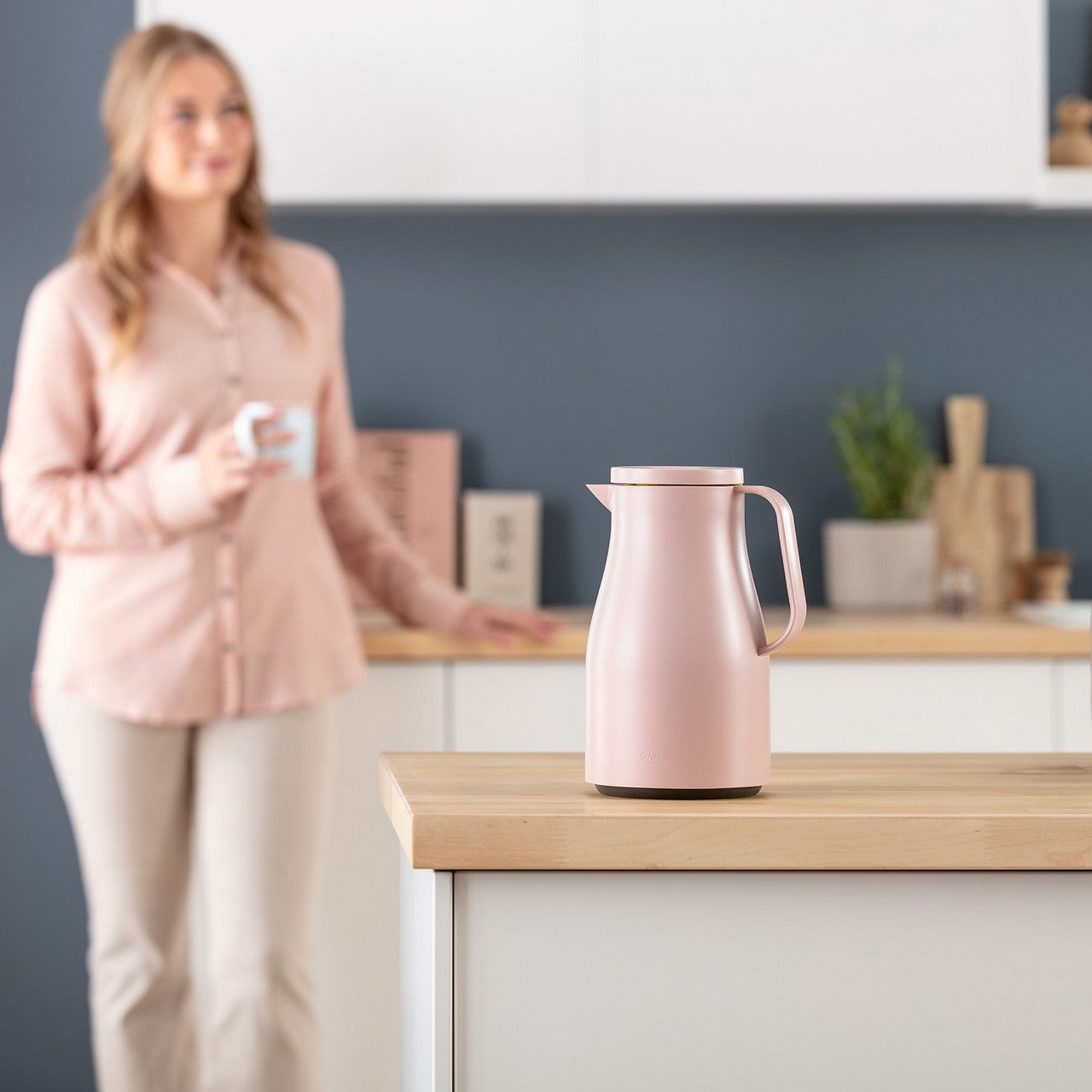 Im Vordergrund steht eine rosafarbene Thermoskanne auf einem Küchentresen. Im Hintergrund ist eine Frau in einem passenden rosa Hemd, die eine Tasse in den Händen hält. Die Küche hat eine moderne, minimalistische Einrichtung.