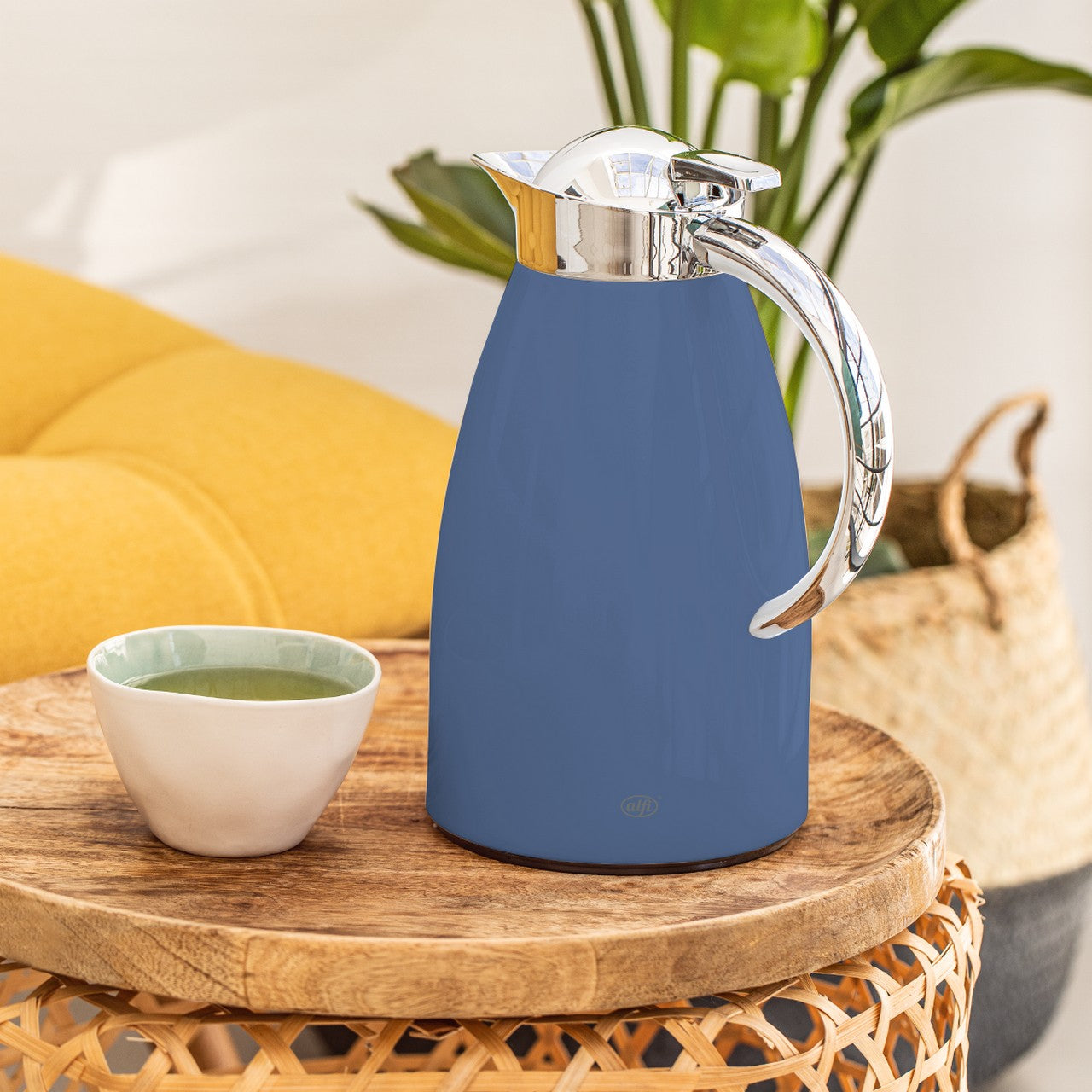 Auf einem runden Holztisch steht eine blaue Thermoskanne mit einem glänzenden Henkel und Deckel. Daneben ist eine kleine, weiße Tasse mit grünem Tee zu sehen. Im Hintergrund befinden sich unscharfe grüne Pflanzen und ein gelbes Kissen.