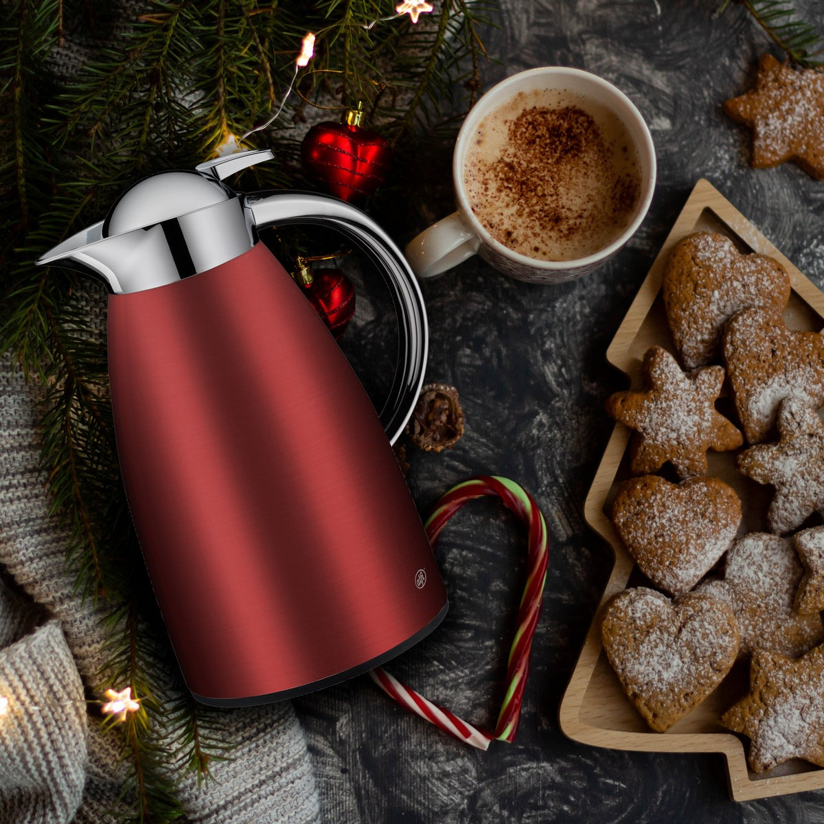 Die Szene zeigt eine rot-silberne Thermoskanne auf einem weihnachtlich dekorierten Tisch mit Tannenzweigen neben einer Kaffeetasse und einem Kekstablett.