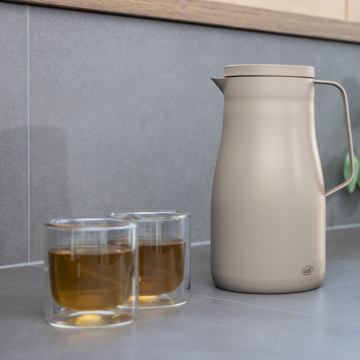 Auf dem Bild steht eine beige Thermoskanne mit Henkel auf einer grauen Arbeitsplatte. Daneben sind zwei doppelwandige Gläser mit bernsteinfarbenem Getränk gefüllt. Im Hintergrund befindet sich eine graue Wand.