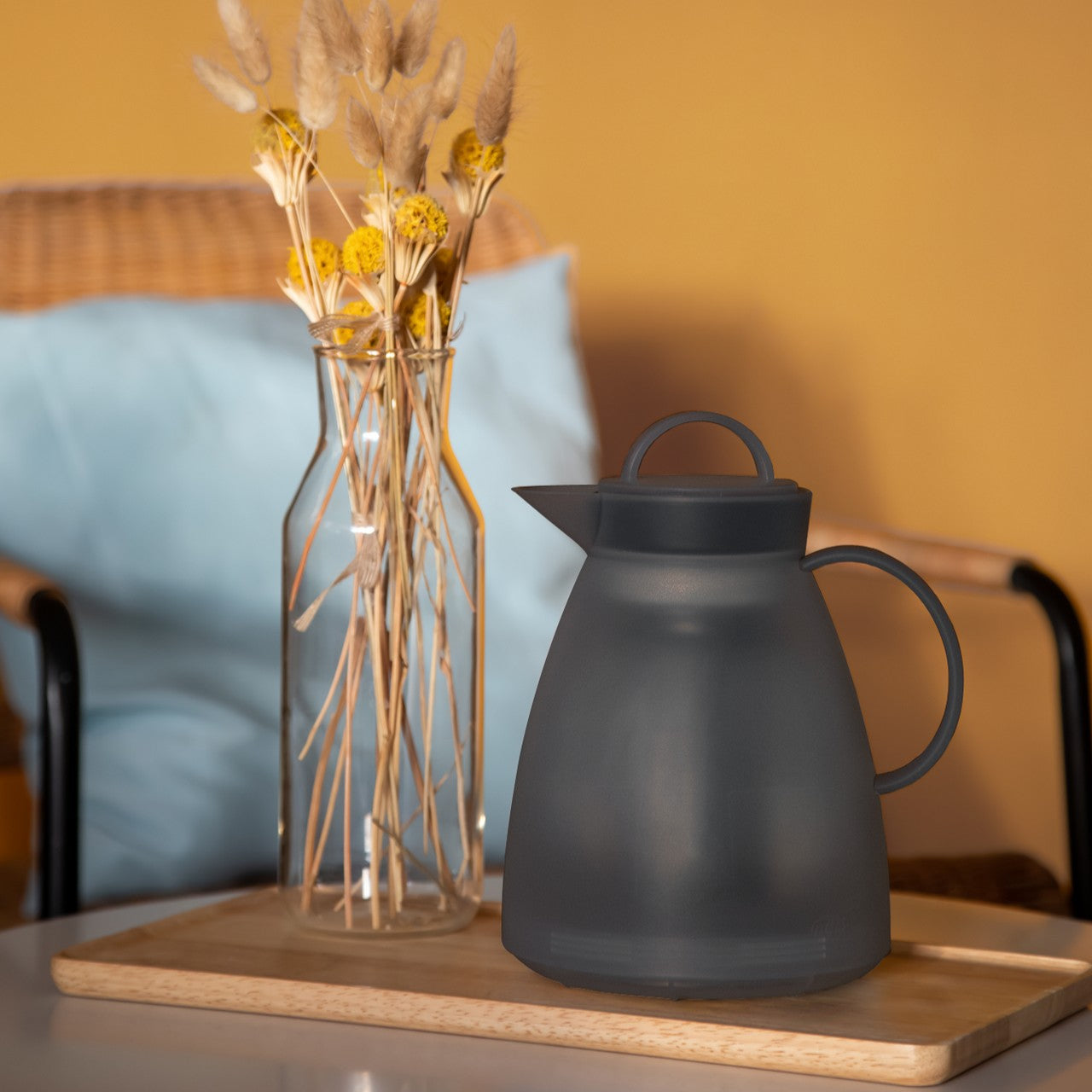 Das Bild zeigt eine grau-matte Teekanne auf einem Holztablett neben einer Glasvase mit getrockneten Gräsern und gelben Blüten. Der Hintergrund ist beige mit einem hellblauen Kissen, das auf einem Stuhl liegt. Die Szene wirkt warm und einladend.