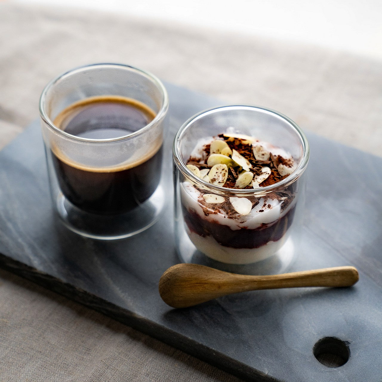 Auf dem Bild stehen zwei durchsichtige Gläser auf einer dunklen Steinplatte: eins mit schwarzem Kaffee und das andere mit einem Schichtdessert, das mit Nüssen und Schokoladenraspeln garniert ist. Daneben liegt ein kleiner Holzlöffel.