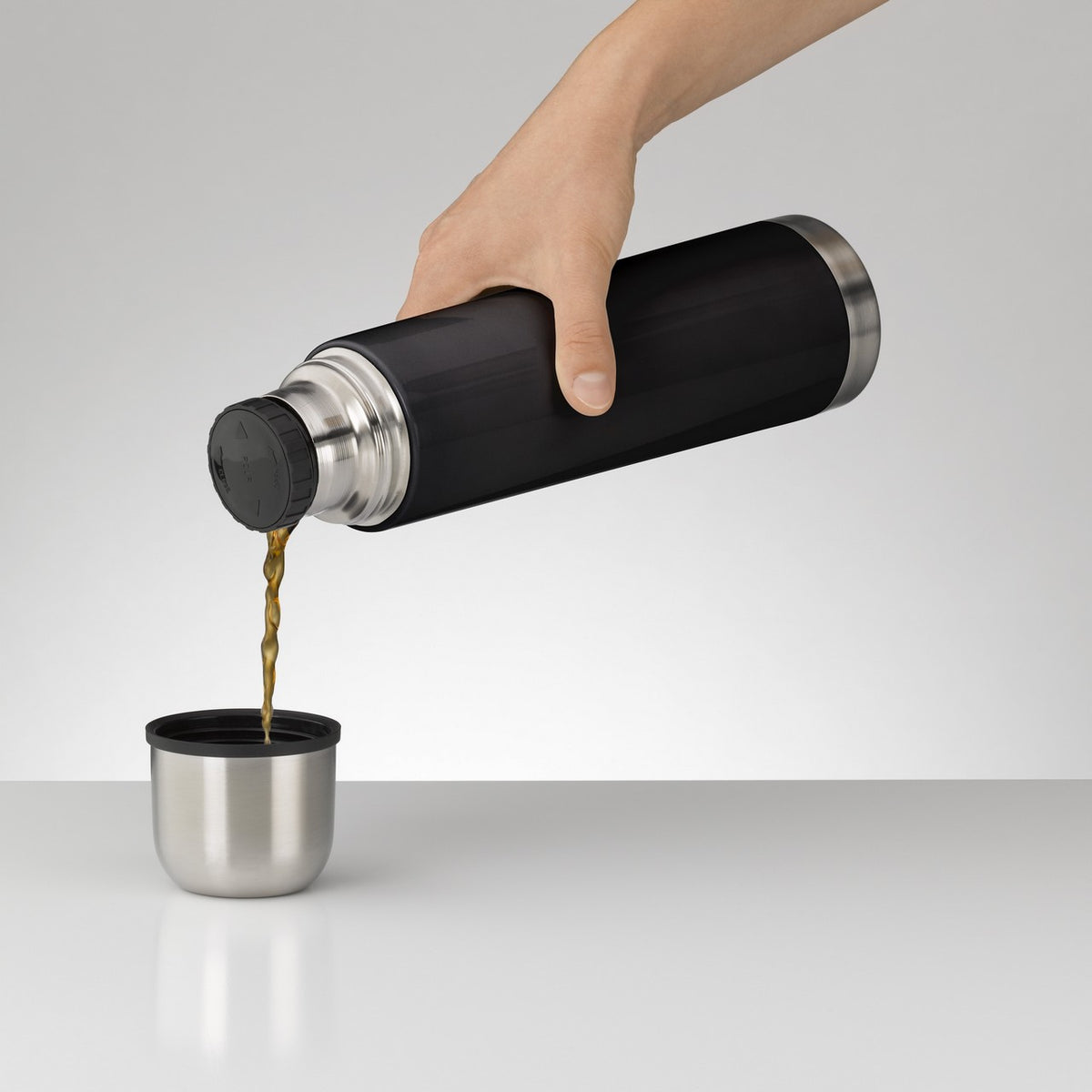 Eine Hand hält eine schwarze Thermoskanne, aus der eine Flüssigkeit in einen kleinen Becher gegossen wird. Der Hintergrund ist schlicht und grau, wodurch der Fokus auf die Kanne und den Becher gerichtet ist.