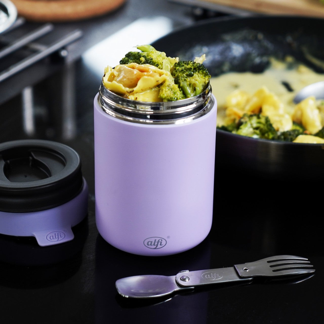Ein lila Thermobehälter ist mit Pasta und Brokkoli gefüllt und steht auf einer dunklen Oberfläche. Daneben liegt ein zusammenklappbares Besteck, und im Hintergrund ist eine Pfanne mit mehr Pasta zu sehen.