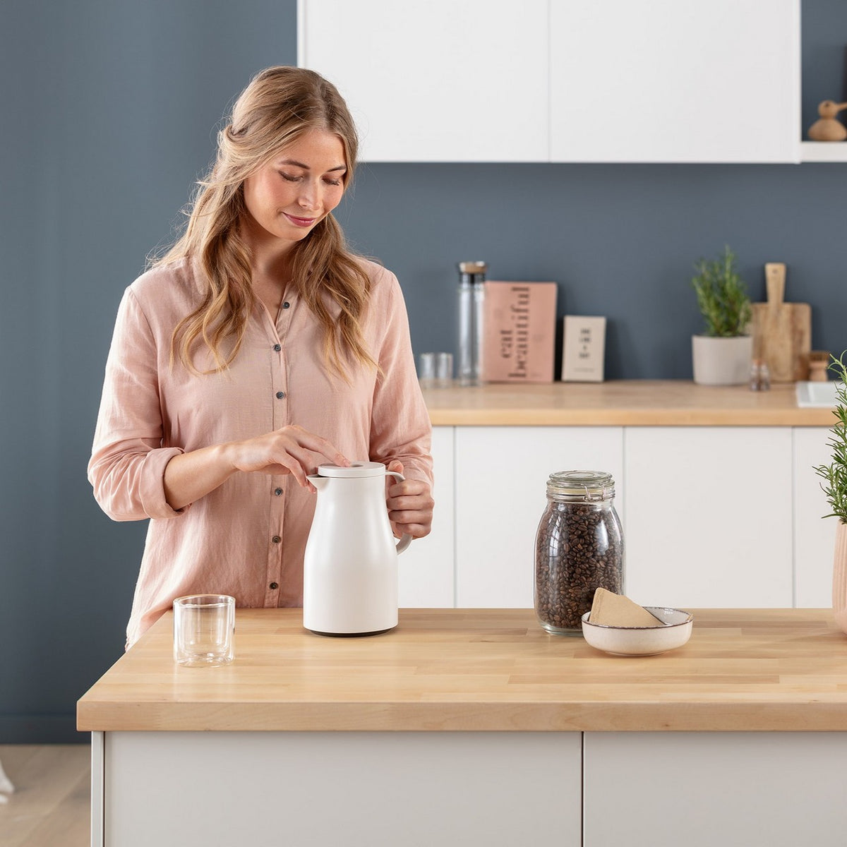 Eine Frau hält lächelnd eine weiße Kanne in einer modernen Küche. Vor ihr stehen ein Glas, ein Glasbehälter mit Kaffeebohnen und eine Schale mit Filtertüten. Die Küche ist schlicht und aufgeräumt mit weißen Schränken.