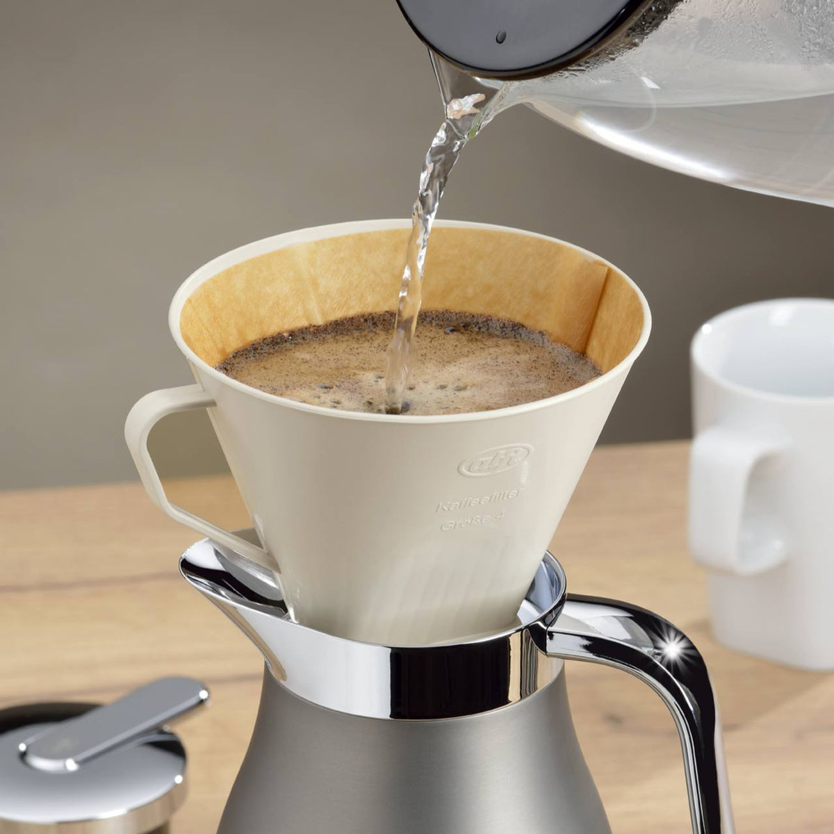 Ein Kaffeebereiter wird mit heißem Wasser über einem Kaffeefilter gefüllt. Darunter steht eine silberne Kanne, im Hintergrund ist eine weiße Tasse.