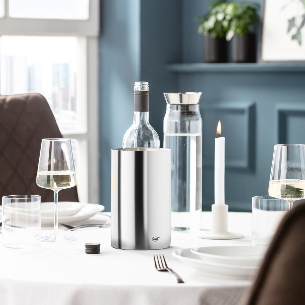 Das Bild zeigt einen elegant gedeckten Tisch mit weißen Kerzen in Kerzenhaltern und einer Edelstahl-Isolierkanne. Im Hintergrund ist eine blaue Wand mit einem Regal, auf dem Pflanzen stehen. Durch das Fenster fällt Tageslicht in den Raum.