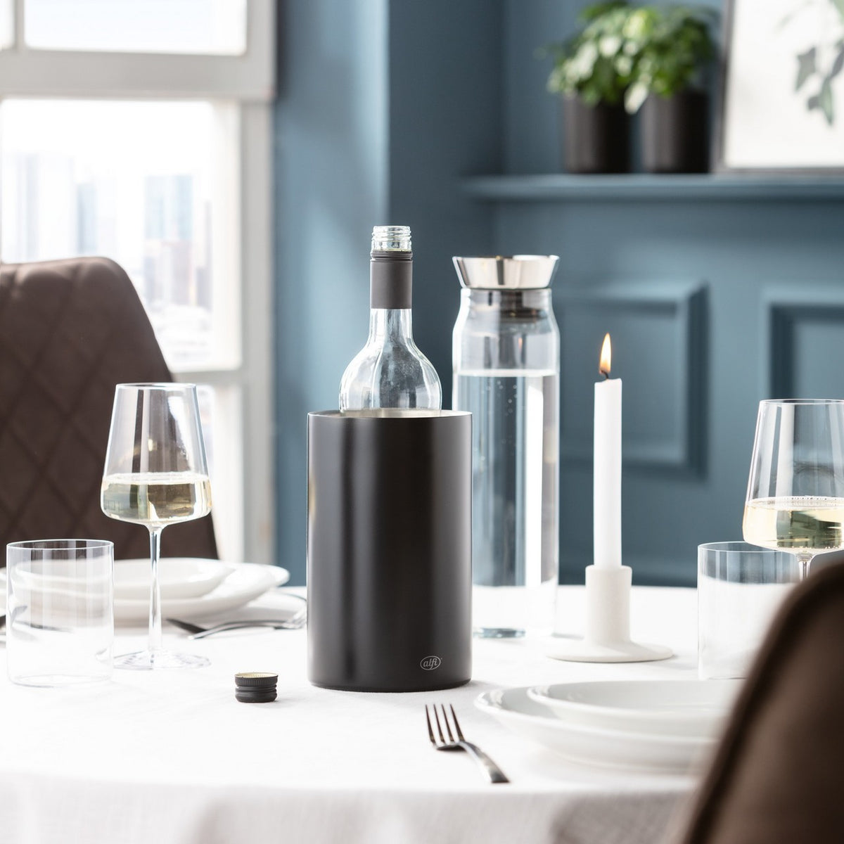 Ein stilvoll gedeckter Tisch mit einer dunklen Wasserflasche und Weingläsern steht im Vordergrund. Eine blaue Wand mit Pflanzen und Dekoration ist im Hintergrund. Der Raum wirkt einladend.