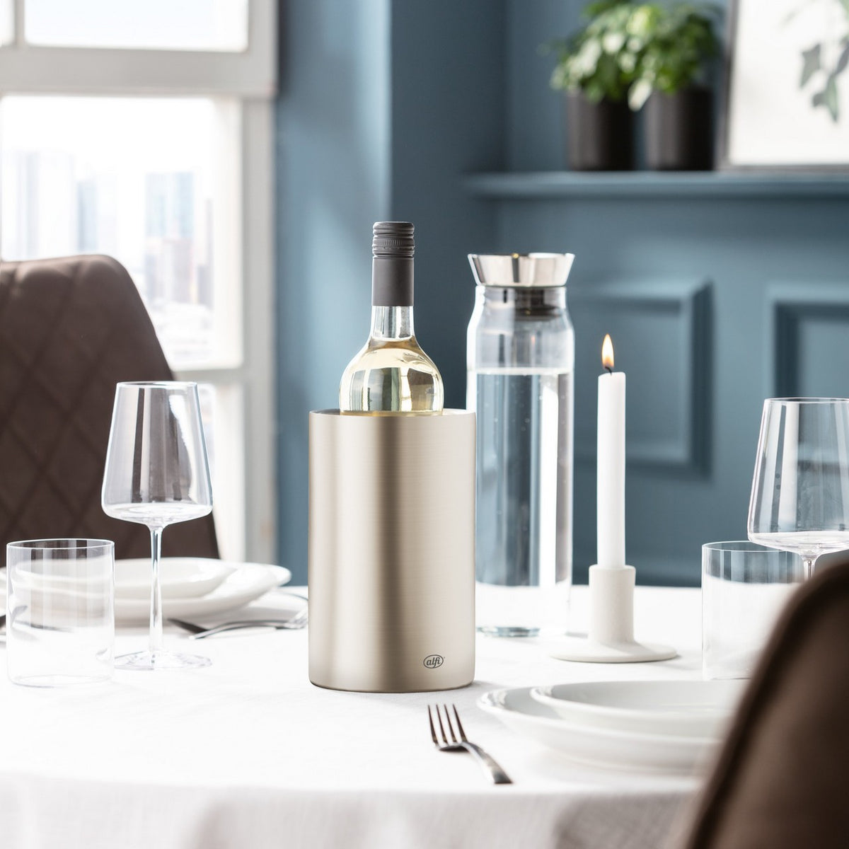 Ein elegant gedeckter Tisch mit Tischtuch, Weingläsern und Kerze steht in einem hellen Raum. Im Hintergrund sind Pflanzen auf einem Regal vor einer blau getäfelten Wand. Tageslicht fällt durch ein Fenster.