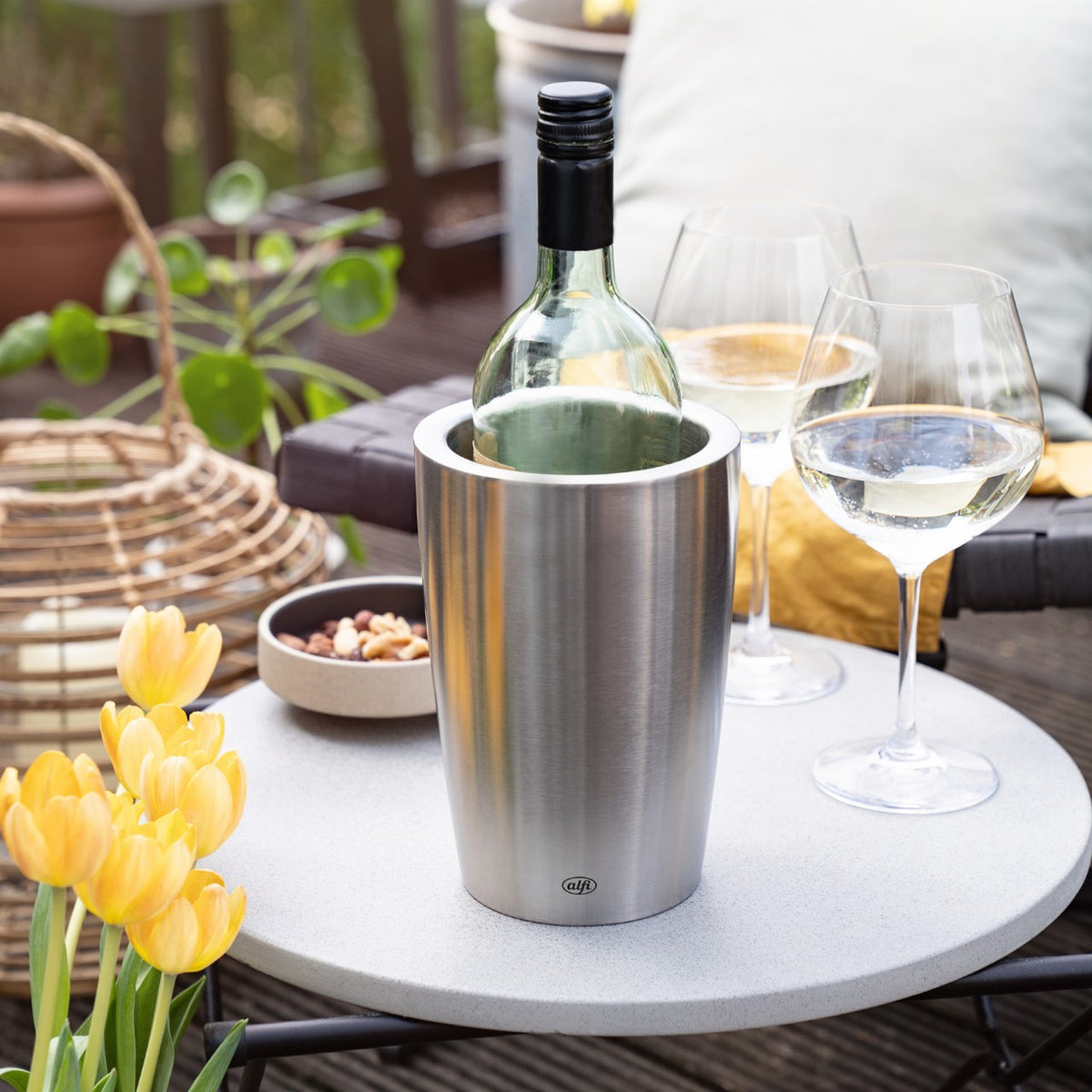 Auf dem Bild steht eine Flasche Wein in einem silbernen Kühler auf einem runden Tisch, daneben zwei Weingläser. Neben dem Tisch sind gelbe Tulpen und eine Schale mit Nüssen zu sehen. Die Szene wirkt wie eine gemütliche Terrasse.