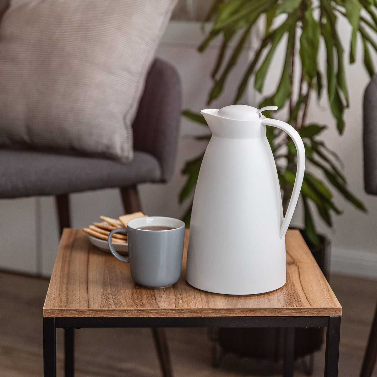 Auf einem kleinen Holztisch stehen eine weiße Kanne und eine graue Tasse mit Flüssigkeit. Daneben liegen einige Kekse auf einem Teller. Im Hintergrund ist ein gemütlicher Raum mit Pflanzen und einem Sessel zu sehen.