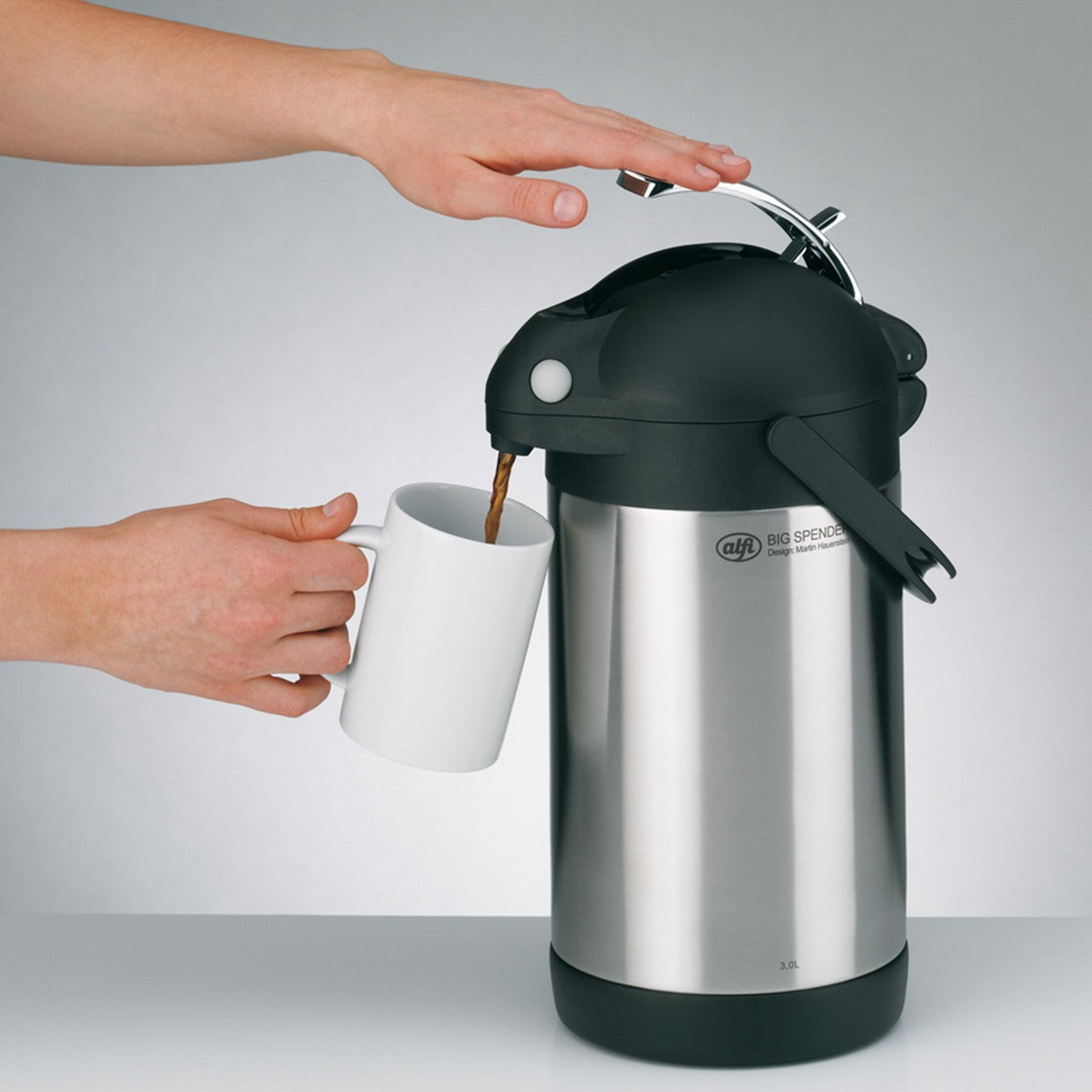 Eine Hand drückt auf die schwarze Oberseite einer silbernen Pumpkanne, während eine andere Hand darunter eine Tasse hält. Die Kanne steht auf einer hellen Oberfläche, und der Hintergrund ist neutral grau.