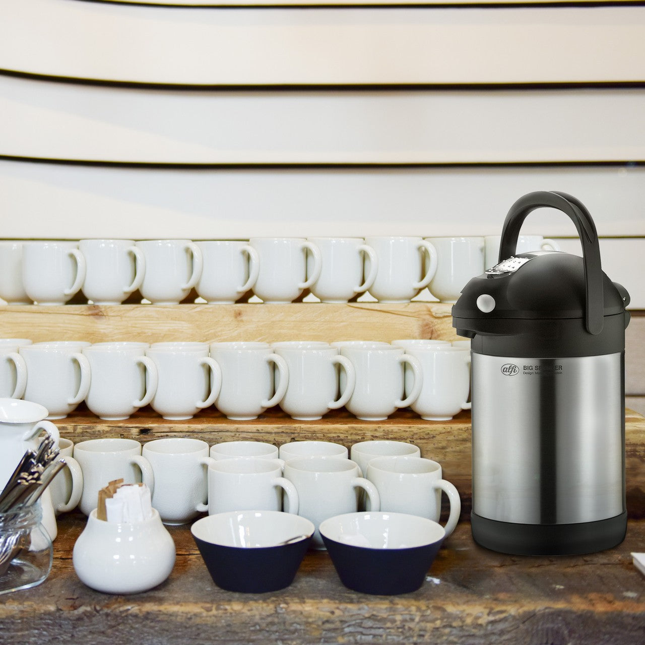 Das Bild zeigt eine Ansammlung von weißen Kaffeetassen, die auf einem Regal gestapelt sind. Daneben steht eine große Thermoskanne aus Edelstahl. Die Anordnung wirkt ordentlich und aufgeräumt.