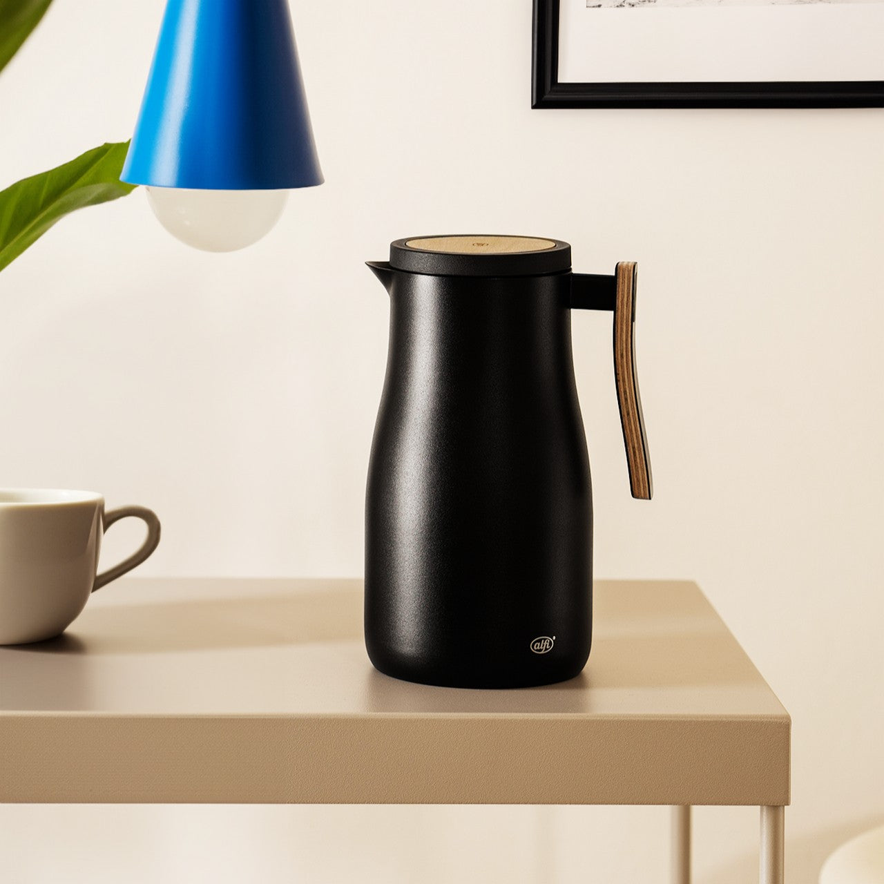 Auf dem Bild ist ein Tisch mit einer schwarzen Thermoskanne und einer kleinen weißen Tasse zu sehen. Im Hintergrund hängen ein blauer Lampenschirm und ein gerahmtes Bild an der Wand. Daneben befindet sich eine grüne Pflanze.