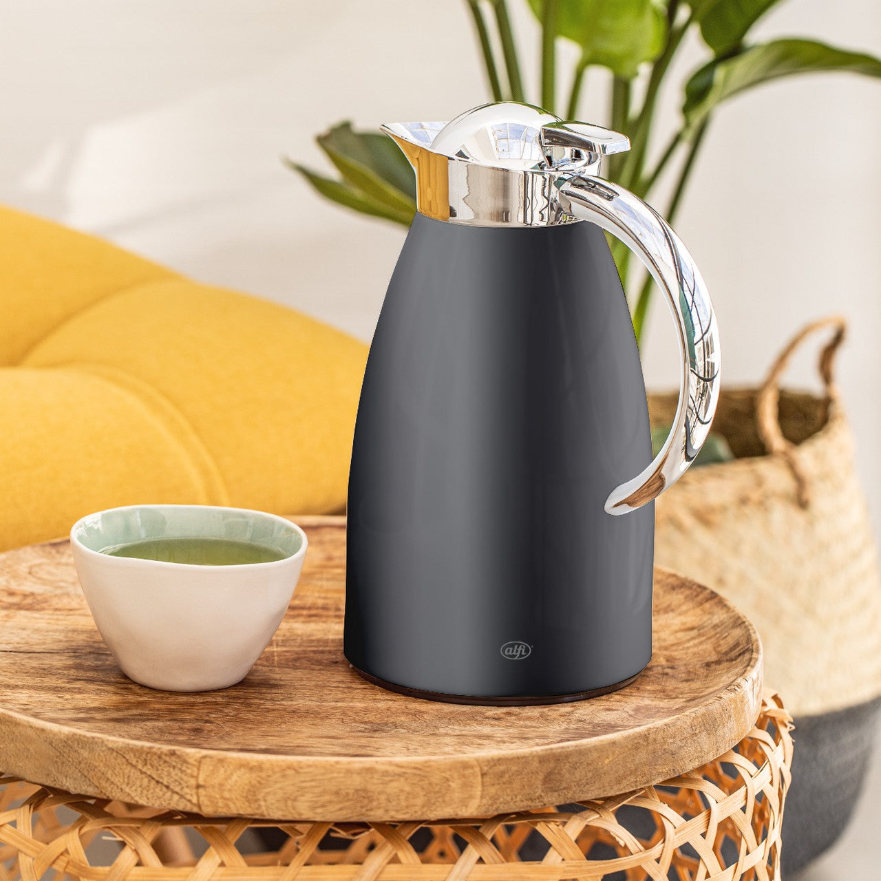 Auf einem runden Holztisch steht eine große, elegante schwarze Thermoskanne mit einem silbernen Deckel und Griff. Neben der Kanne befindet sich eine kleine, weiße Tasse mit grünem Inhalt. Im Hintergrund sieht man ein gelbes Kissen und eine Pflanze.