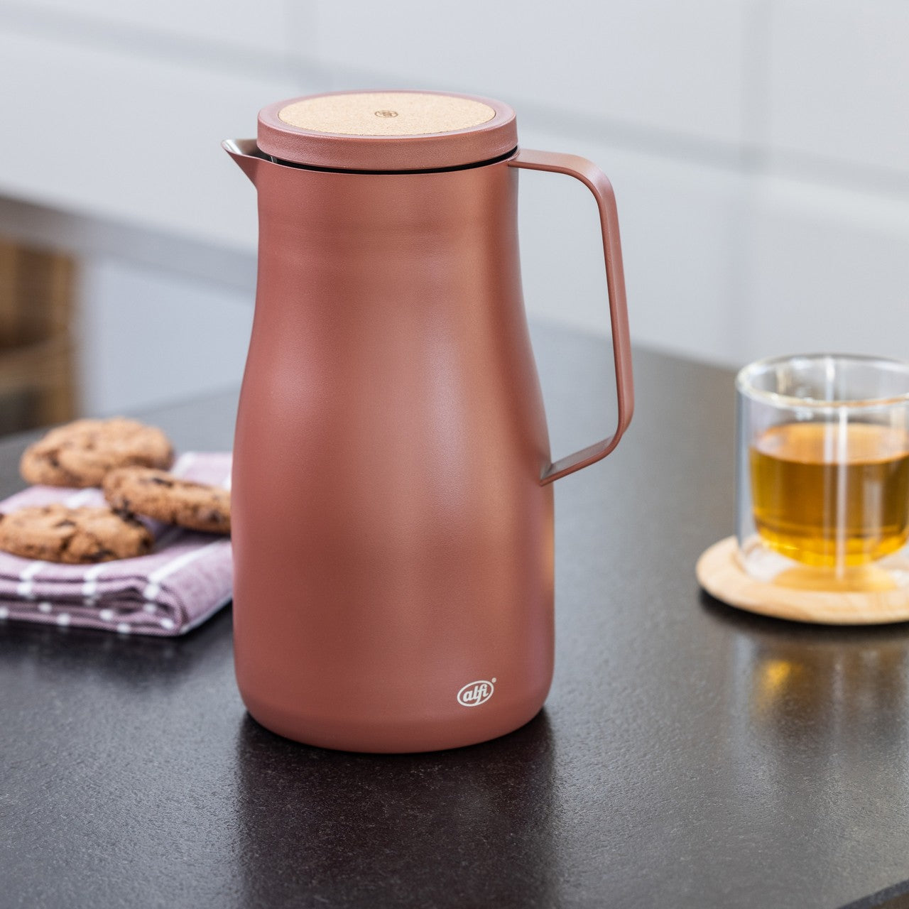 Auf dem Bild ist eine braune Thermoskanne aus Metall mit einem Holzdeckel zu sehen, die auf einem Tisch steht. Daneben liegen Kekse auf einem gefalteten Tuch, und ein Glas Tee steht auf einem Untersetzer. Die Szene wirkt gemütlich und einladend.