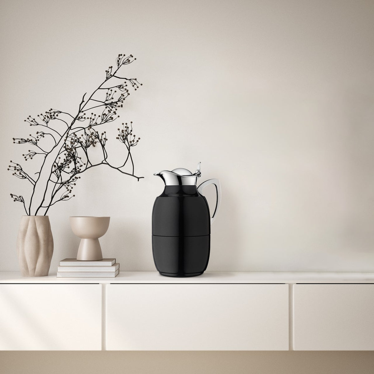 Eine schwarze Isolierkanne steht auf einem hellen Sideboard neben drei gestapelten Büchern. Links davon sind eine Vase mit Zweig und ein Becher. Der Hintergrund ist minimalistisch beige.