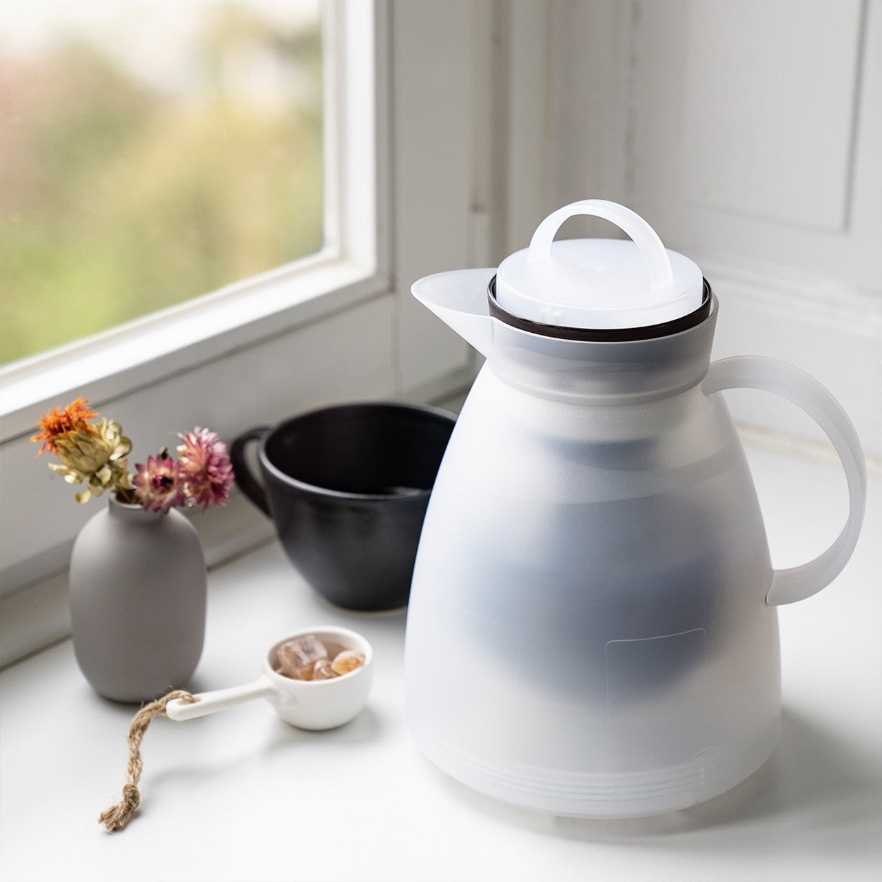 Auf dem Bild sieht man einen weißen Wasserkocher auf einer Fensterbank. Daneben stehen eine schwarze Tasse, ein Gefäß mit Zuckerwürfeln und eine Vase mit getrockneten Blumen. Im Hintergrund ist ein Fenster mit unscharfen Pflanzen.