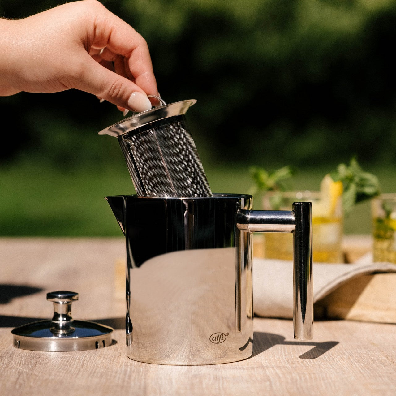 Eine Hand hält einen metallenen Teefilter über eine glänzende, silberne Teekanne auf einem Holztisch. Der Deckel der Teekanne liegt daneben und im Hintergrund sind verschwommen Pflanzen und eine mit Getränken gefüllte Karaffe zu sehen.
