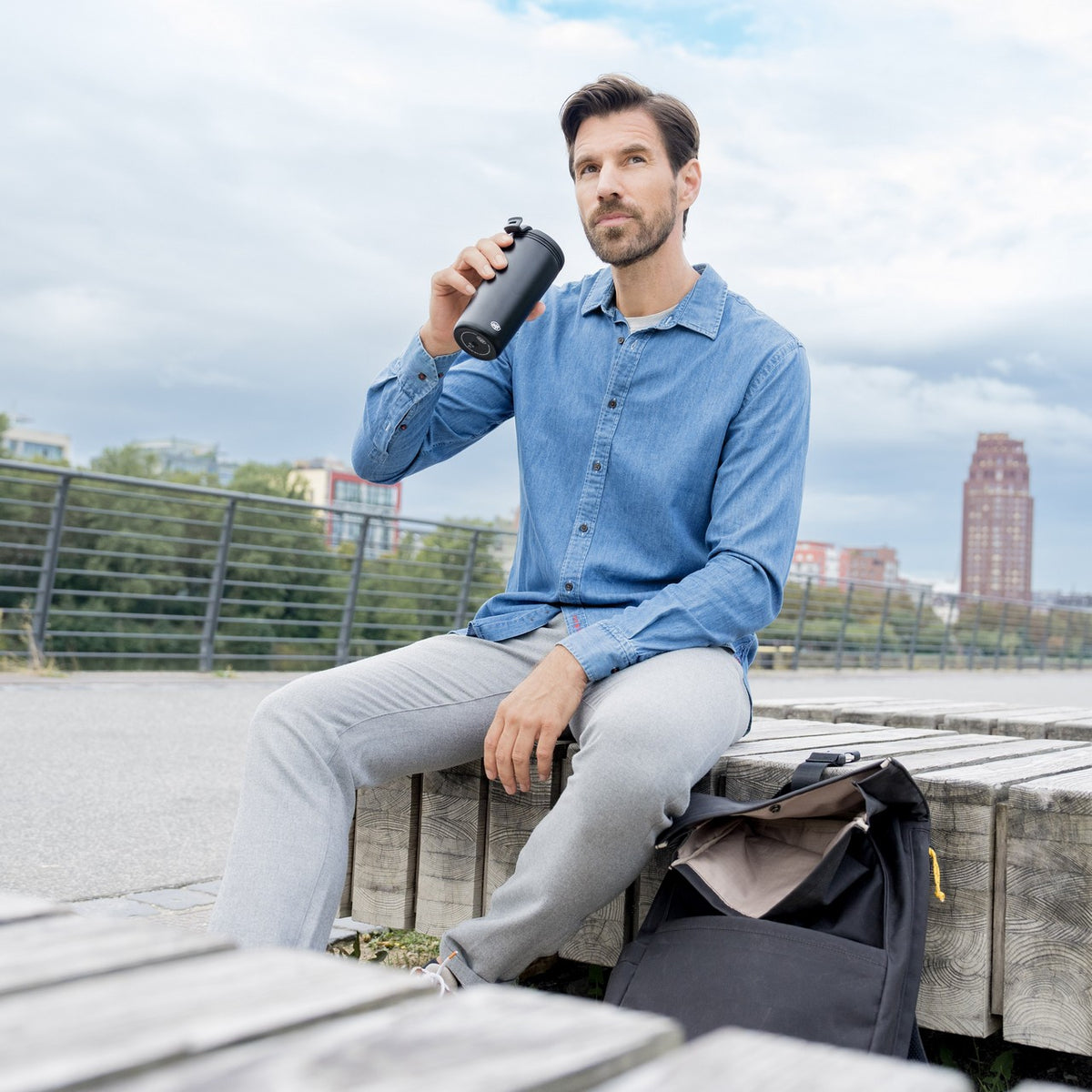 Ein Mann sitzt entspannt auf einem hölzernen Steg am Wasser und trinkt aus einer Wasserflasche. Hinter ihm sind Bäume und moderne Gebäude zu sehen. Neben ihm liegt eine schwarze Tasche.