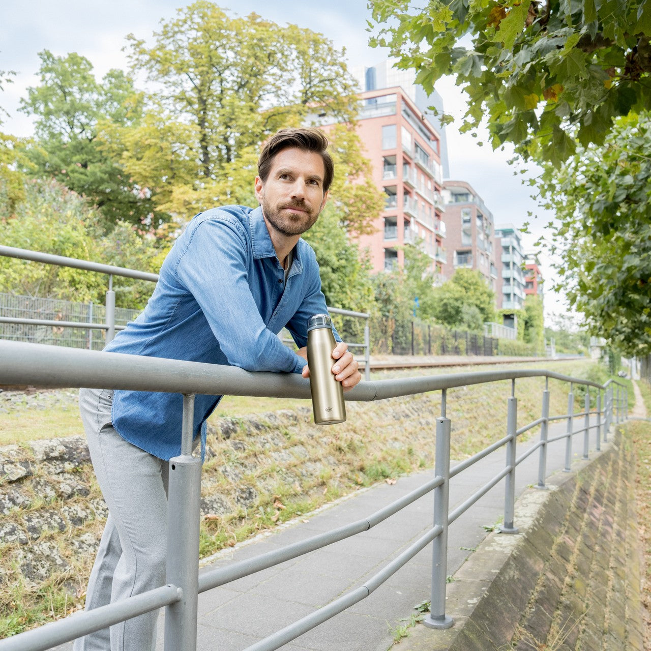 Ein Mann in einem blauen Hemd lehnt sich freundlich lächelnd an ein Geländer. Im Hintergrund sind eine Wiese und einige Bäume sowie mehrstöckige Häuser zu sehen. Die Szene wirkt hell und freundlich.