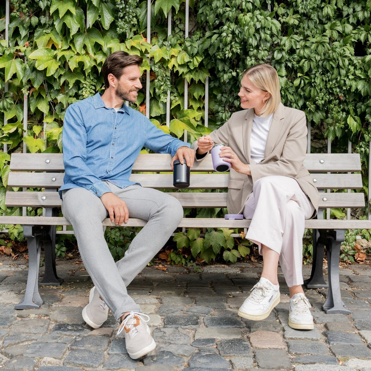 Auf dem Bild sitzen ein Mann und eine Frau auf einer Parkbank vor einer grünen, mit Efeu bewachsenen Wand. Sie lächeln sich an und halten in ihren Händen je einen Becher. Beide wirken entspannt und es scheint, als ob sie sich angeregt unterhalten.